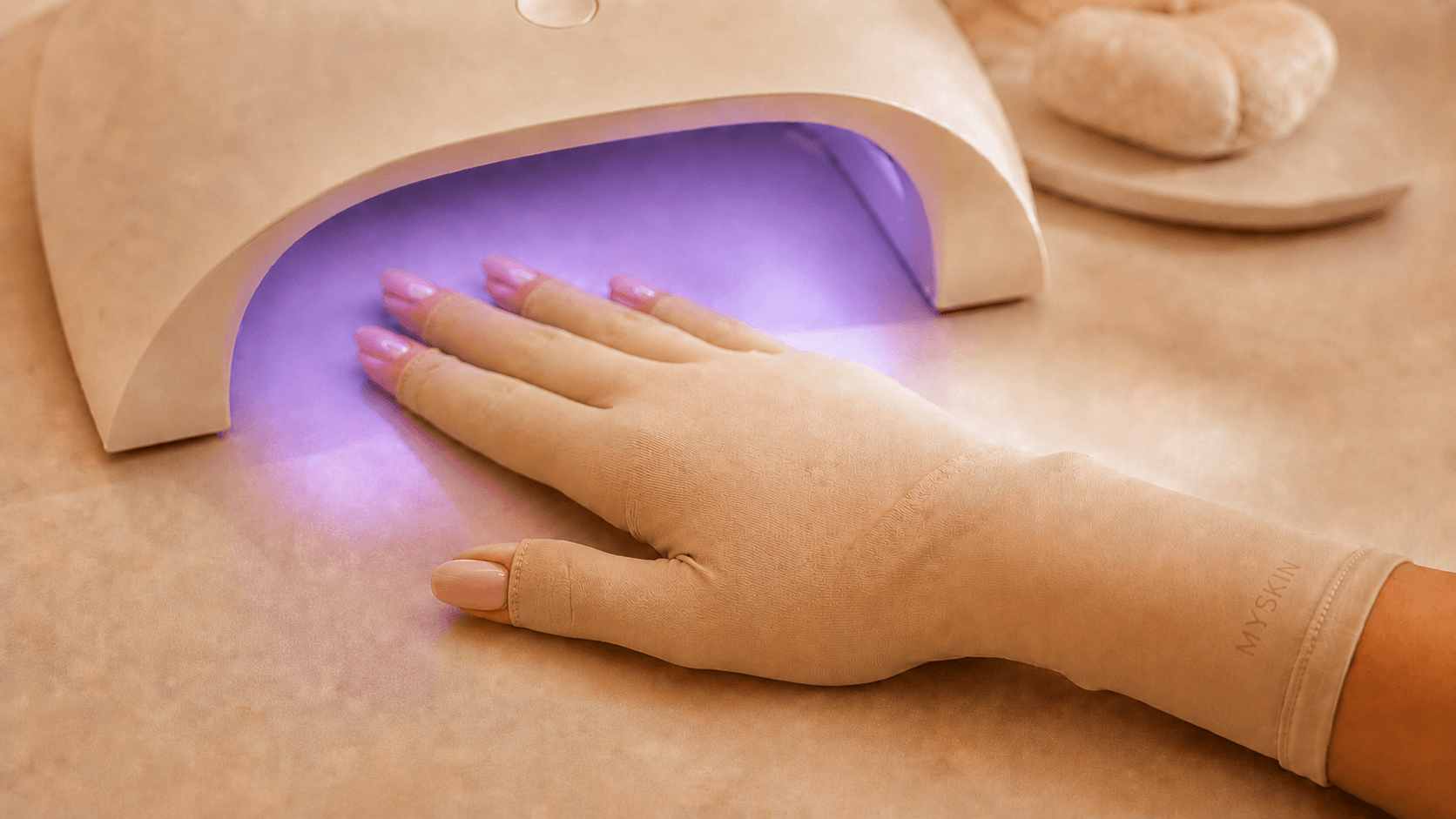 Hand wearing a nude MySkin UV-protective glove beneath an illuminated manicure lamp, with the nails left exposed